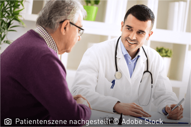 Freundlich lächelnder junger Arzt schaut seinen älteren Patienten aufmunternd an: Mastozytose ist nicht heilbar, die Symptome lassen sich aber lindern.